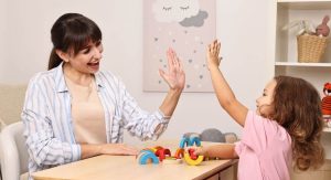 Dorosła kobieta i kilkuletnia dziewczynka siedzą naprzeciwko siebie. Kobieta unosi dłoń w geście przywitania.